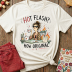 An off-white graphic t-shirt with a humorous illustration of a woman having a hot flash while drinking tea, styled in a flat-lay on a wooden background with red jeans and a floral card.