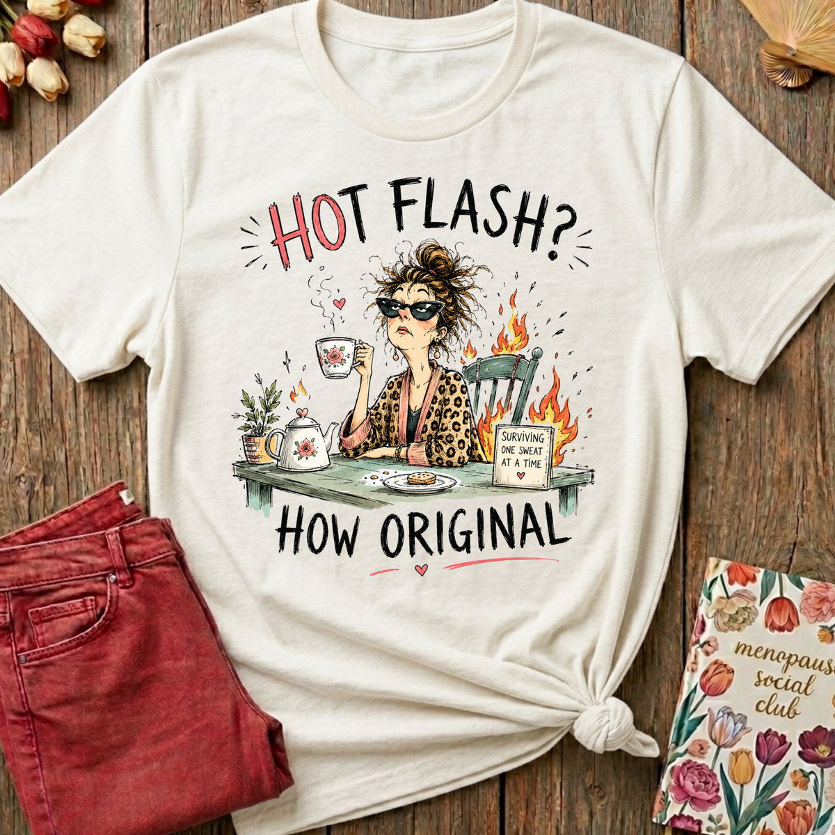 An off-white graphic t-shirt with a humorous illustration of a woman having a hot flash while drinking tea, styled in a flat-lay on a wooden background with red jeans and a floral card.