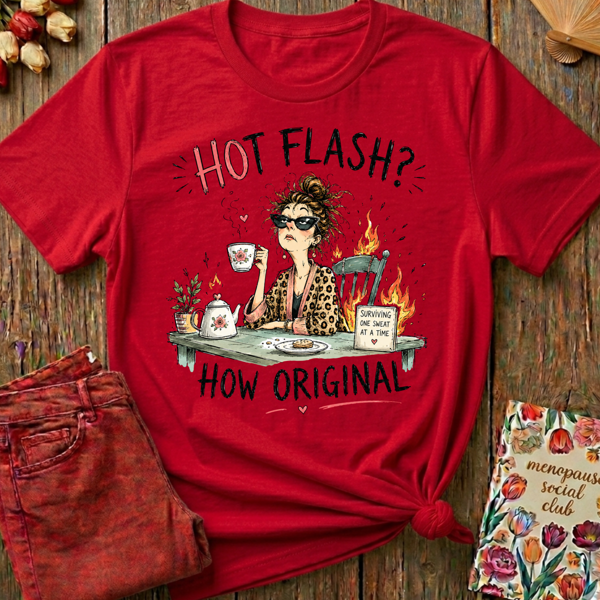 A red graphic t-shirt featuring a cartoon illustration of a woman in a leopard print top and sunglasses, holding a mug with flames, surrounded by the text 'HOT FLASH? HOW ORIGINAL'. The shirt is styled in a flat-lay on a rustic wooden background with a pair of distressed red jeans and a floral book.